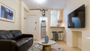 A living room with a black leather sofa, a round coffee table, a TV mounted on the wall, and a kitchen area in the background.