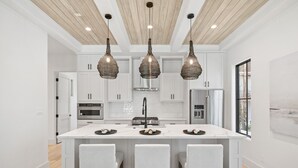 Una cocina moderna con armarios blancos, una isla central con taburetes de bar, iluminación colgante y una campana extractora de acero inoxidable.