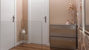 Une pièce avec un plancher en bois, une porte blanche, une commode à tiroirs, un vase contenant des fleurs séchées et un miroir accroché au mur.