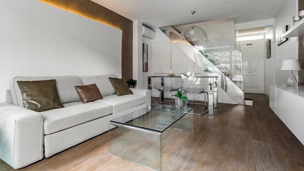 Un salon moderne avec une table basse en verre, un canapé blanc et un escalier menant à un balcon.