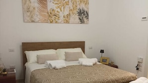 A double bed with a patterned bedspread, multiple pillows, and a headboard. There is a nightstand with a lamp and a small decorative item. A large abstract painting is on the wall above the bed. - Cornflower House (San Benedetto del Tronto)