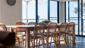 Un coin repas avec une table en bois et des chaises en osier, de grandes fenêtres et une horloge au mur.