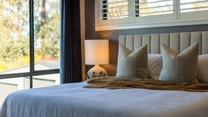 Un lit bien fait, avec une tête de lit, une lampe de chevet et une vue sur les arbres à travers une fenêtre munie de stores.