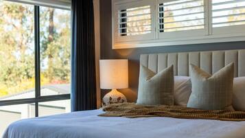 A neatly made bed with a headboard, a bedside lamp, and a view of trees through a window with blinds.