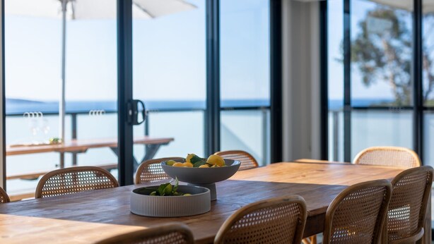 Un coin repas avec une table en bois et des chaises en osier, de grandes fenêtres donnant sur la mer et un saladier rempli de fruits posé sur la table.