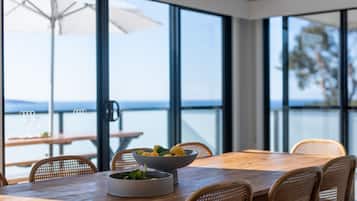 A dining area with a wooden table and wicker chairs, large windows with a view of the sea, and a bowl of fruit on the table.
