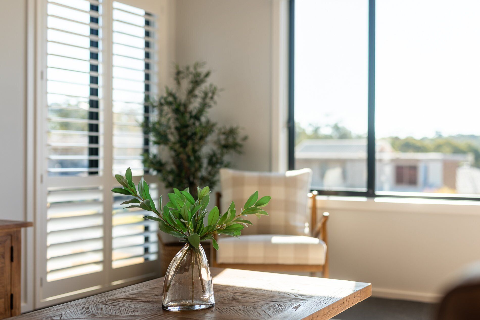 A clear view of a room with a wooden table, a glass vase with green foliage, a chair, and a window with blinds.