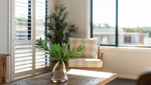 On voit clairement une pièce avec une table en bois, un vase en verre contenant des feuilles vertes, une chaise et une fenêtre avec des stores.