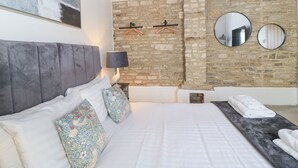 A bedroom with a stone wall, a bed with white bedding, a grey headboard, and two wall-mounted mirrors. - Mayfield Cottage (Malmesbury)