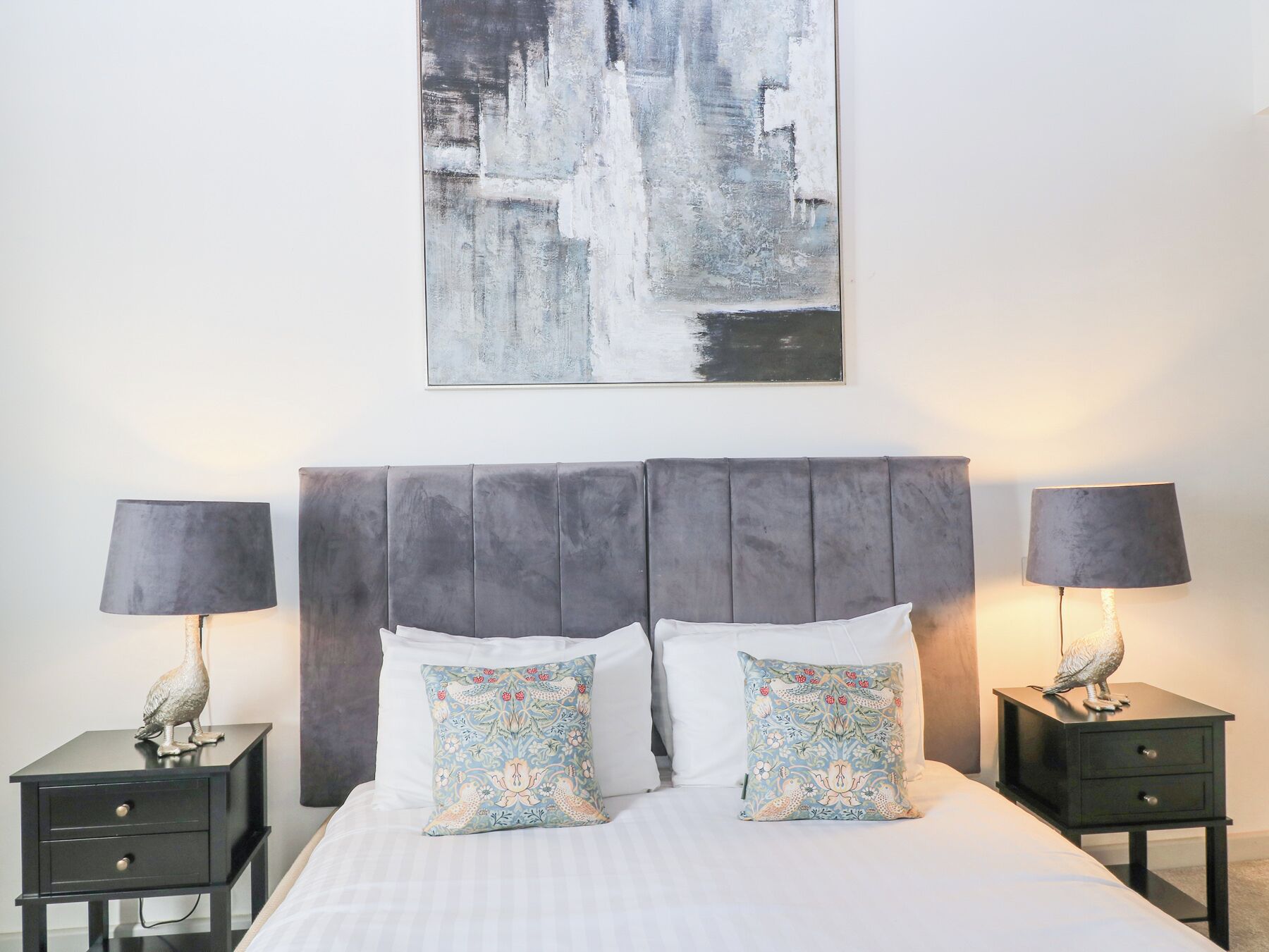 Una cama bien hecha con un cabecero gris, dos mesitas de noche con lámparas y almohadas decorativas.