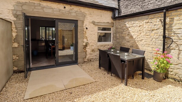 A stone building with a glass door leading to an interior space, a patio with a table and chairs, and a gravel ground. - Mayfield Cottage (Malmesbury)