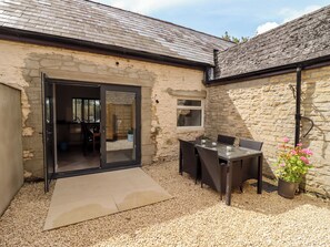 A stone building with a glass door leading to an interior space, a patio with a table and chairs, and a gravel ground. - Mayfield Cottage (Malmesbury)