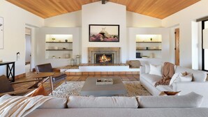 Un salón amplio con techo de madera, una chimenea y un cuadro grande encima. Hay dos sofás, una mesa de centro y un televisor sobre un mueble.