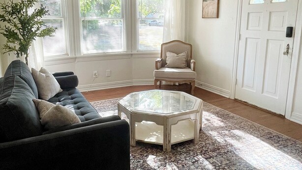 Ein Wohnzimmer mit einem dunklen Sofa, einem Glastisch, einem weißen Sessel und einem gemusterten Teppich.