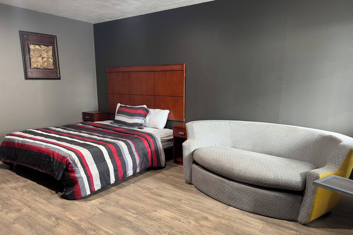 Hotellihuone, jossa on sänky, tuoli, yöpöytä ja seinällä oleva kehystetty kuva.