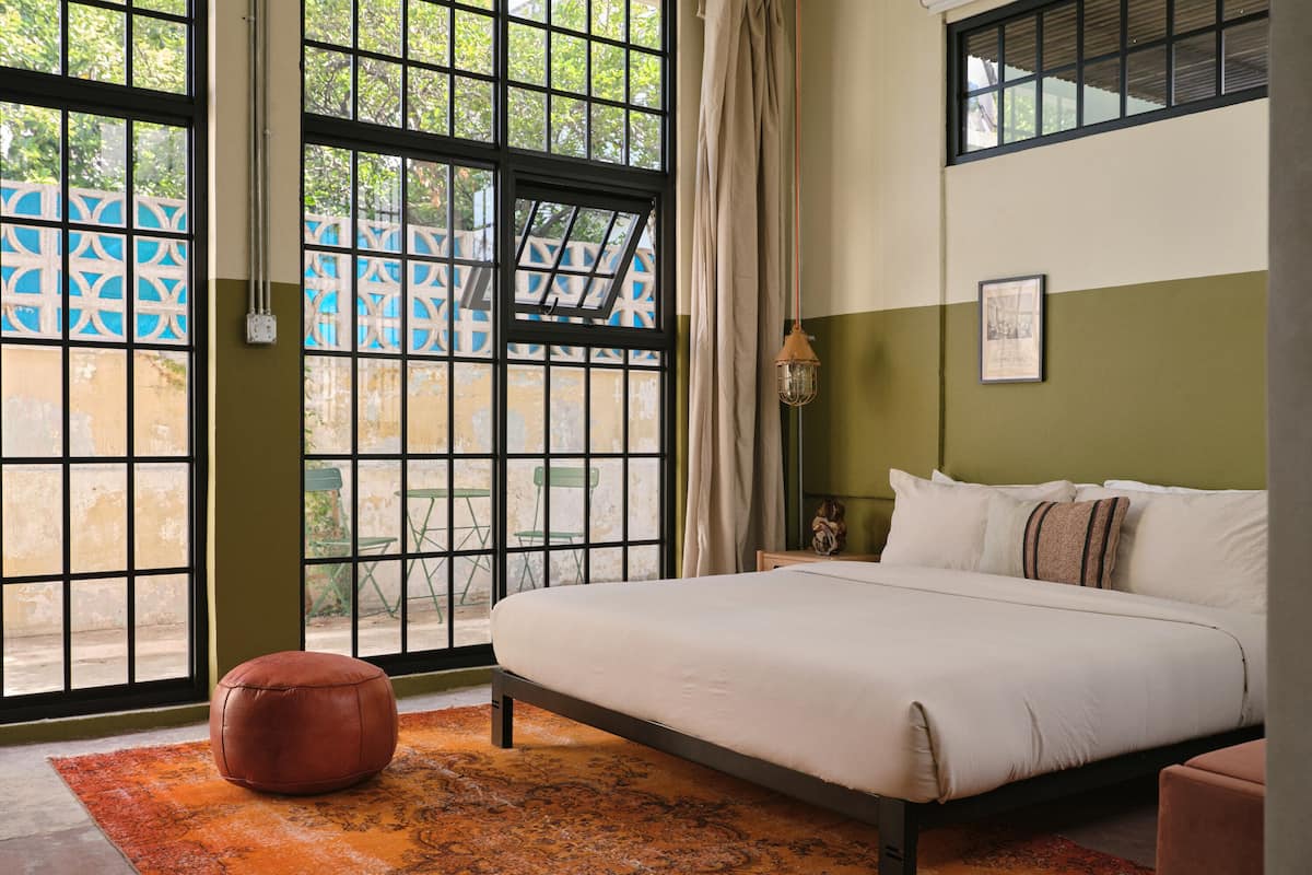 Une chambre avec un grand lit, une fenêtre donnant sur un paysage verdoyant, un tapis à motifs et une petite table avec une lampe.