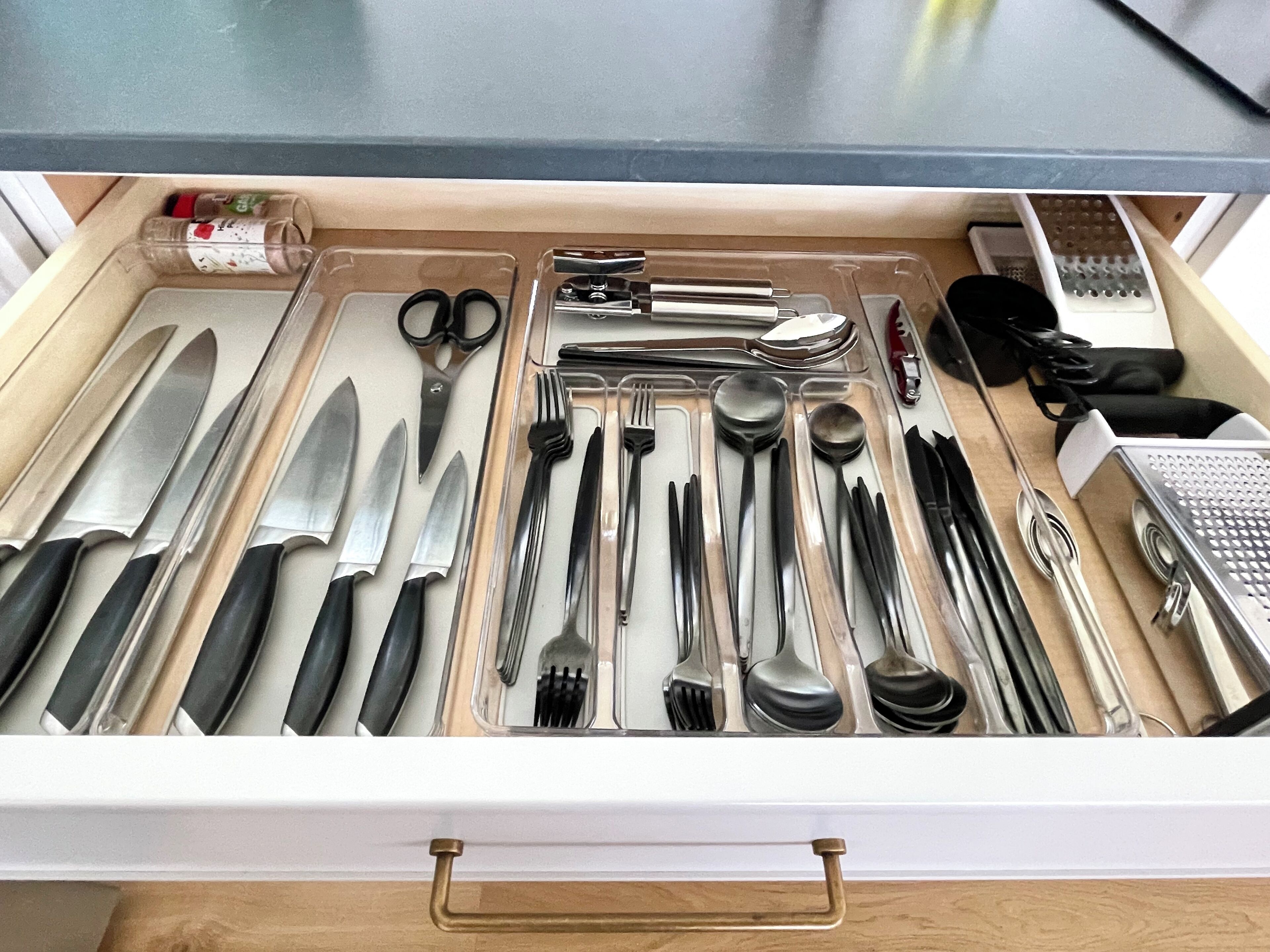 Eine Schublade mit verschiedenen Küchenutensilien, darunter Messer, Gabeln, Löffel und eine Reibe.