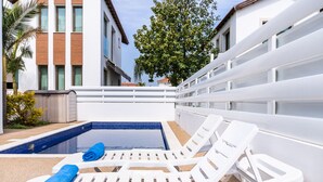 A pool area with white lounge chairs and a blue towel, surrounded by a white fence and buildings.