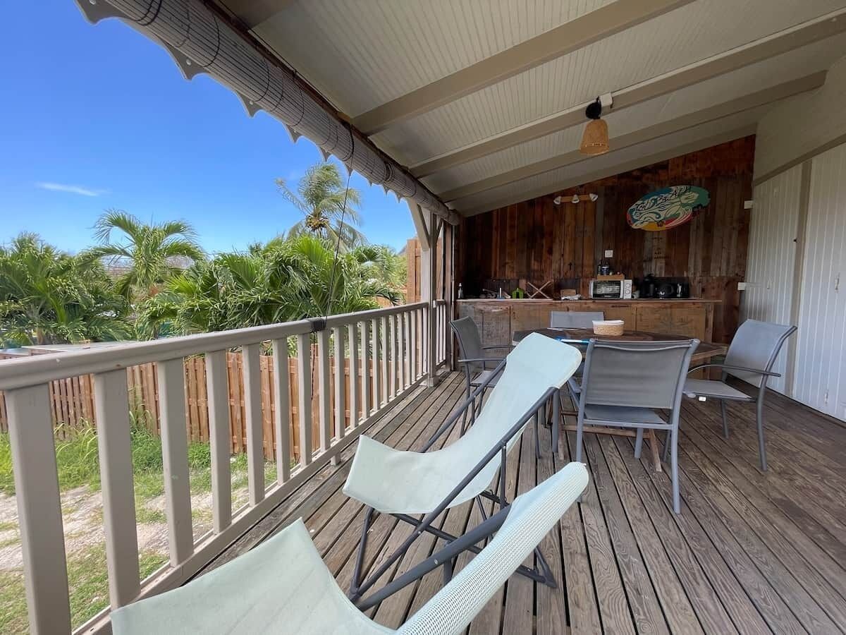 A covered outdoor area with a wooden deck, lounge chairs, a table, and a bar area with kitchen appliances.