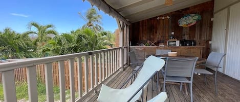 A covered outdoor area with a wooden deck, lounge chairs, a table, and a bar area with kitchen appliances.