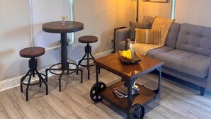 A living room with a grey sofa, a coffee table with a tray of lemons, a small round table with stools, and an air conditioning unit on the wall. - Tiny home in Watauga. “Casita”  Beautiful Cottage! (Watauga)