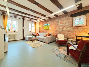 Ein gemütliches Wohnzimmer mit freiliegenden Holzbalken, einer Steinwand, einem Sofa, einem roten Sessel und einem kleinen Tisch mit einer Vase und Weingläsern.