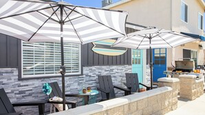 Un patio al aire libre con sombrillas a rayas, sillas negras, un muro de piedra y una parrilla.
