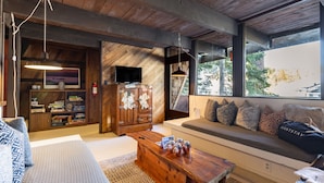 A cozy living room with a wooden ceiling, a large window with a view, a sofa with patterned cushions, and a coffee table with jars.