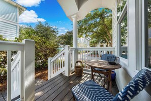 A porch with a wooden table and blue patterned chairs, surrounded by white railings and a view of trees and houses.