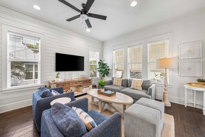 A spacious living room with a ceiling fan, a flat-screen TV, and a large window with blinds.