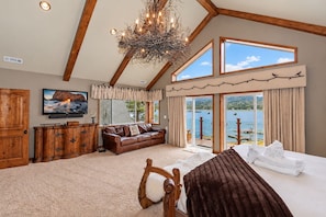 A bedroom with a large bed, a wooden dresser, a leather sofa, a television, and a view of a lake through large windows.