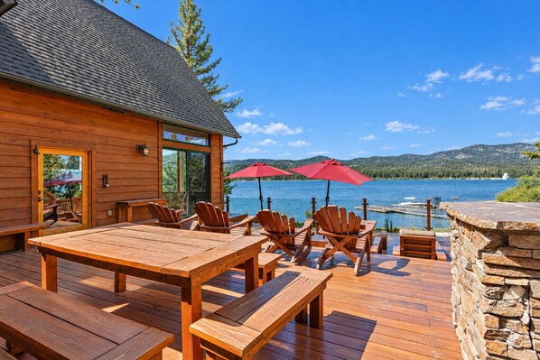 A wooden deck with a table and chairs, overlooking a lake with mountains in the background.