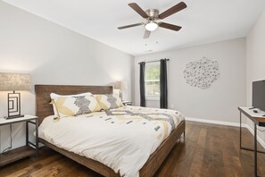 Une chambre avec un lit en bois, un ventilateur de plafond, une œuvre d’art murale, une fenêtre avec des rideaux et un meuble pour la télévision.