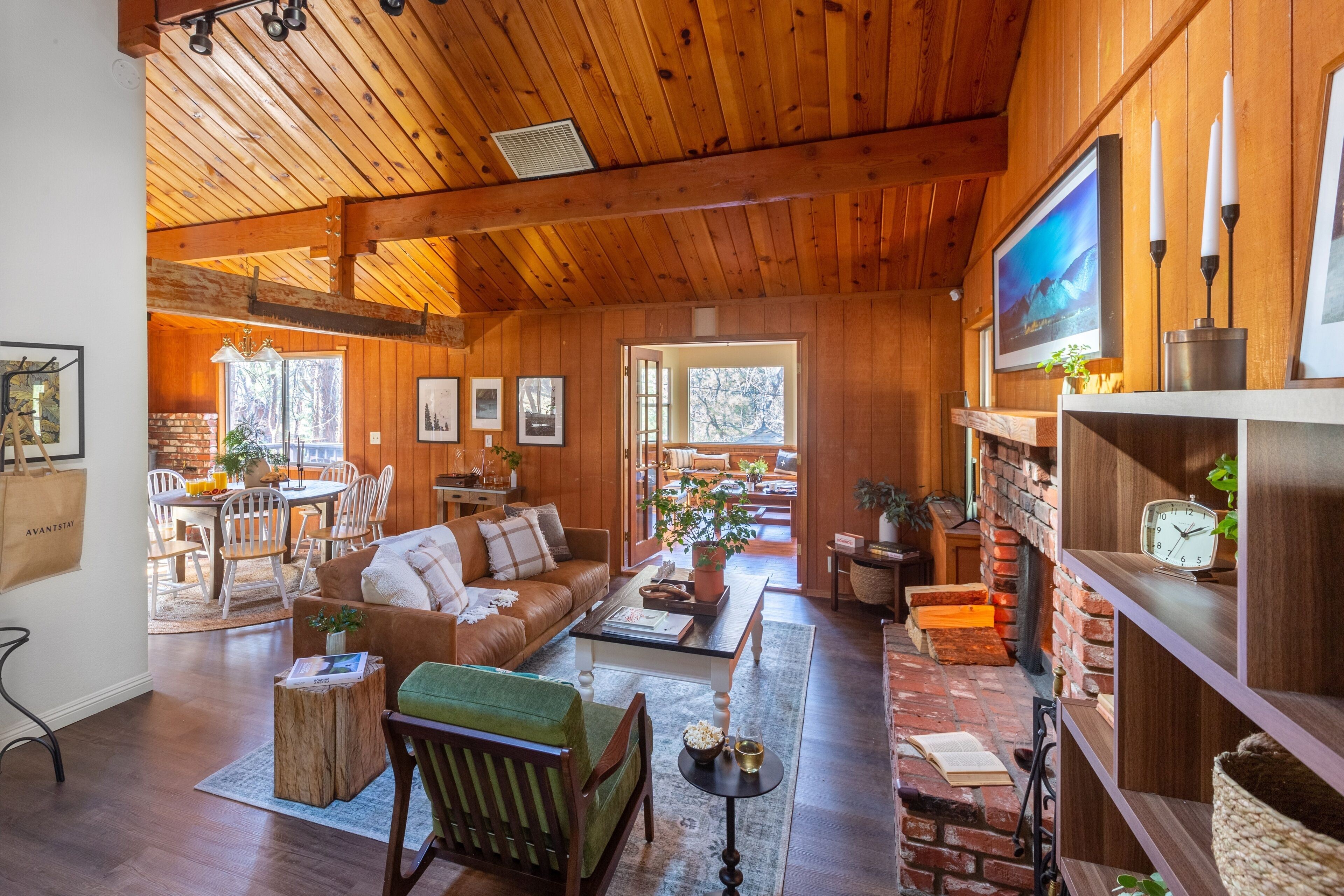 A cozy cabin interior with a wooden ceiling, a dining area, a living room with a sofa and coffee table, and a bookshelf.