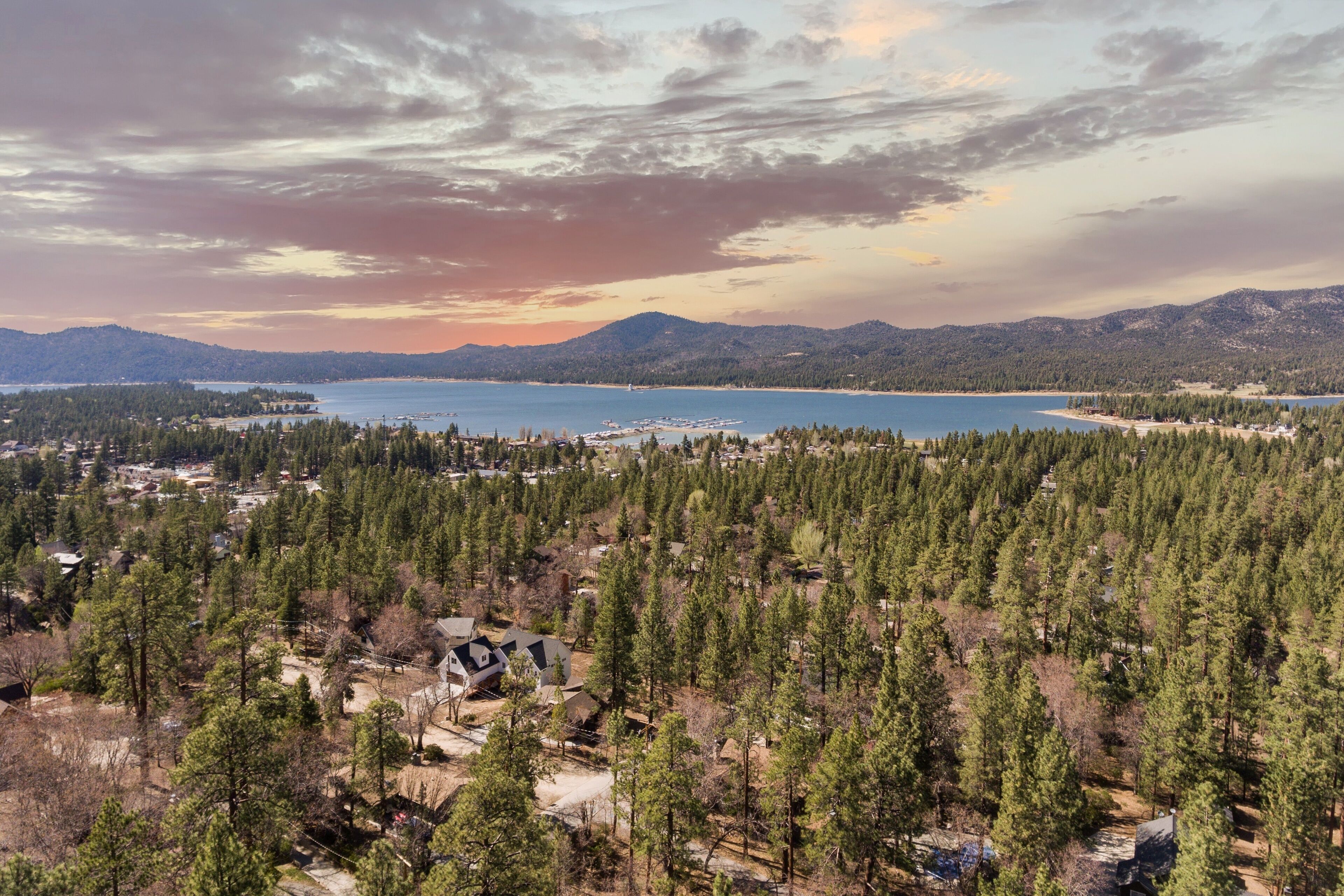 Et boligområde med huse omgivet af tæt skov, et stort vandområde og bjerge i baggrunden.