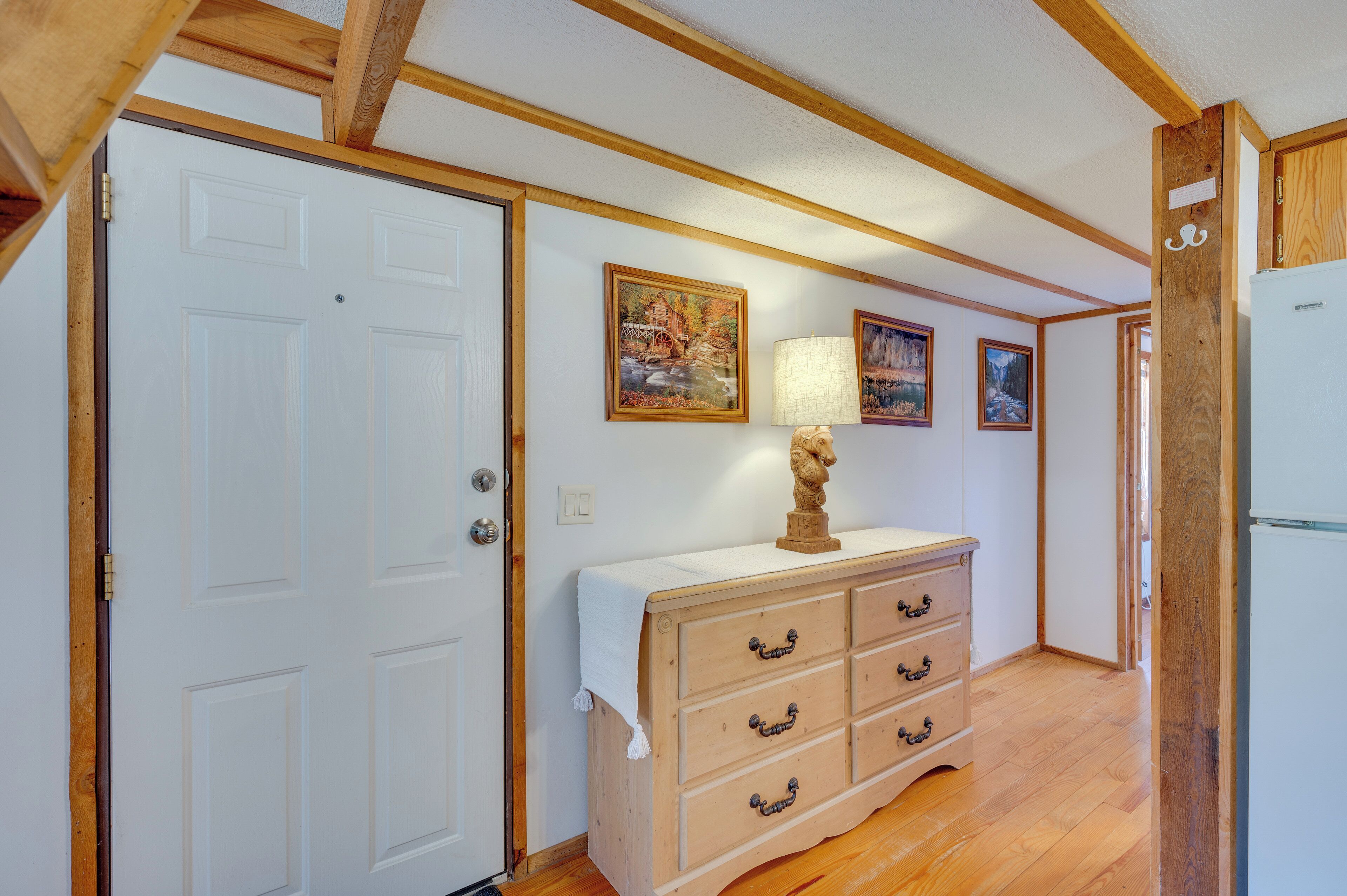 A hallway with wooden flooring, a white door, a wooden dresser with a lamp, and two framed pictures on the wall.