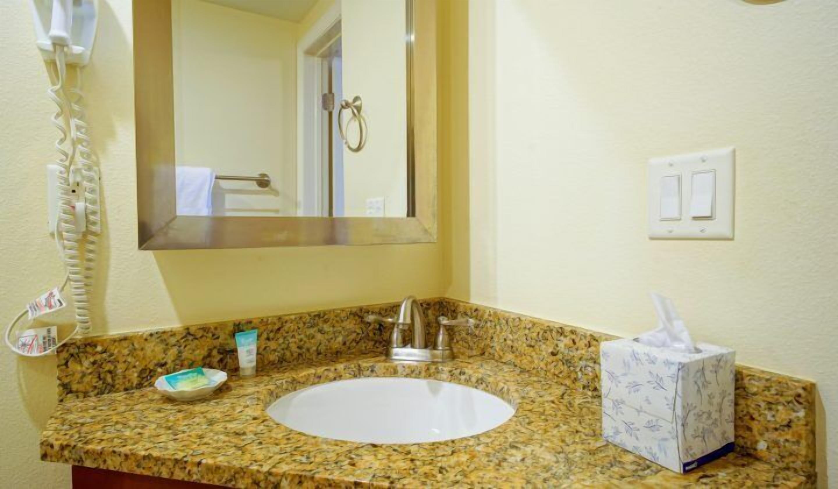 Un baño con encimera de granito, un lavabo, un espejo, un secador de pelo y una caja de pañuelos.