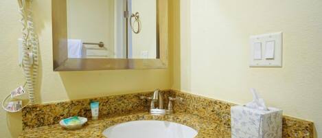 Un baño con encimera de granito, un lavabo, un espejo, un secador de pelo y una caja de pañuelos.