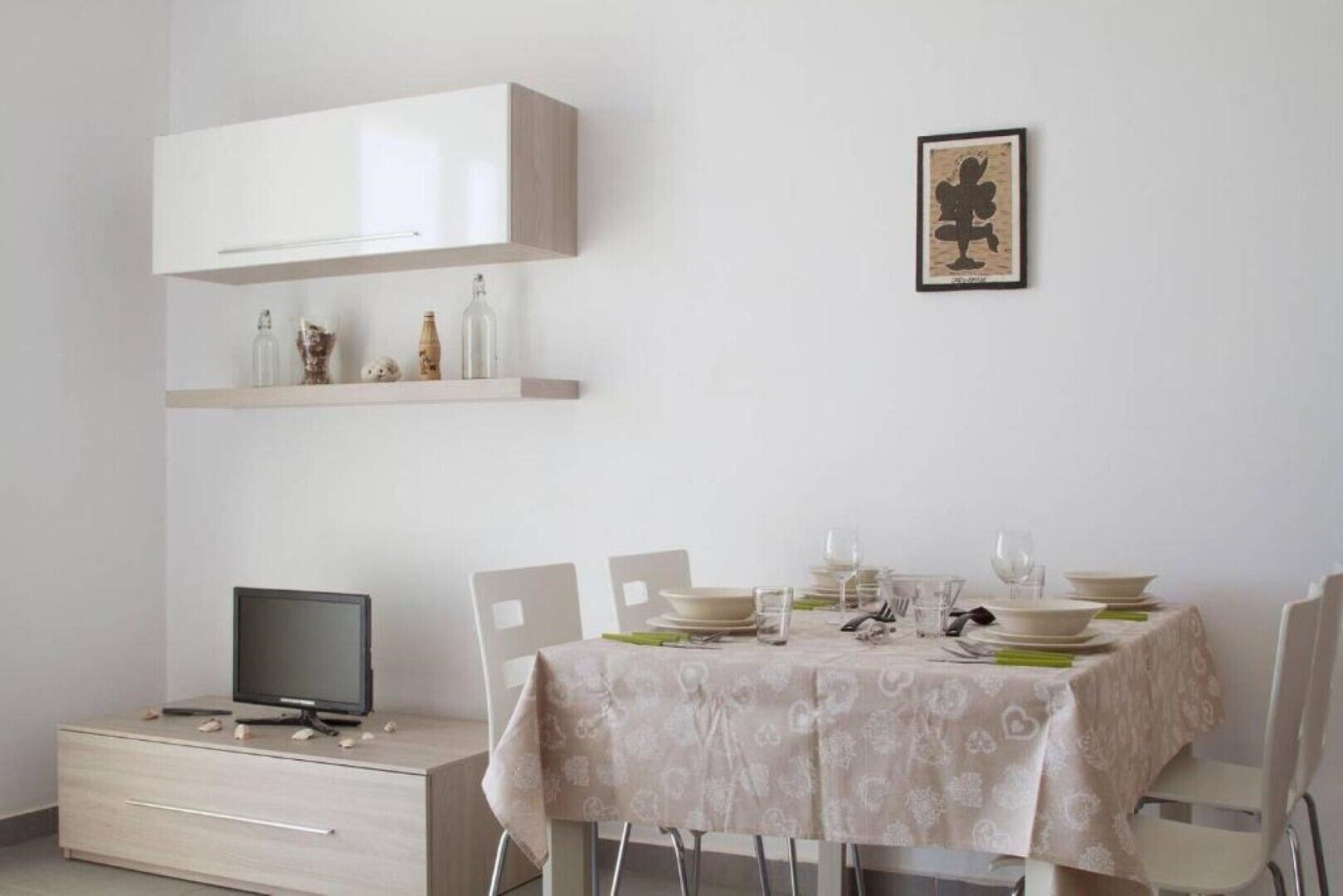 A dining area with a table set for four, a television on a stand, and a shelf with decorative items.