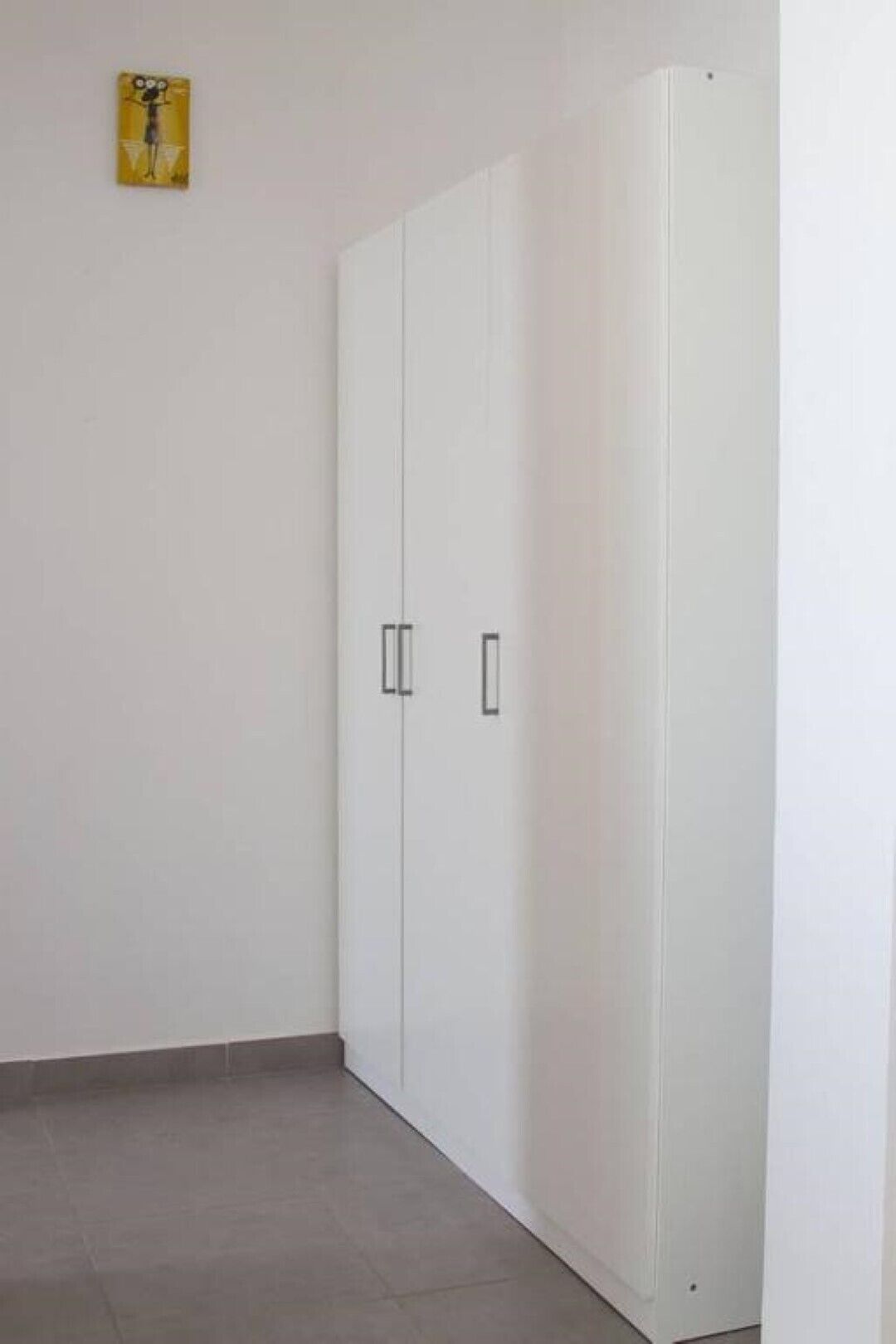 A room with a white wardrobe, a tiled floor, and a small yellow artwork on the wall.