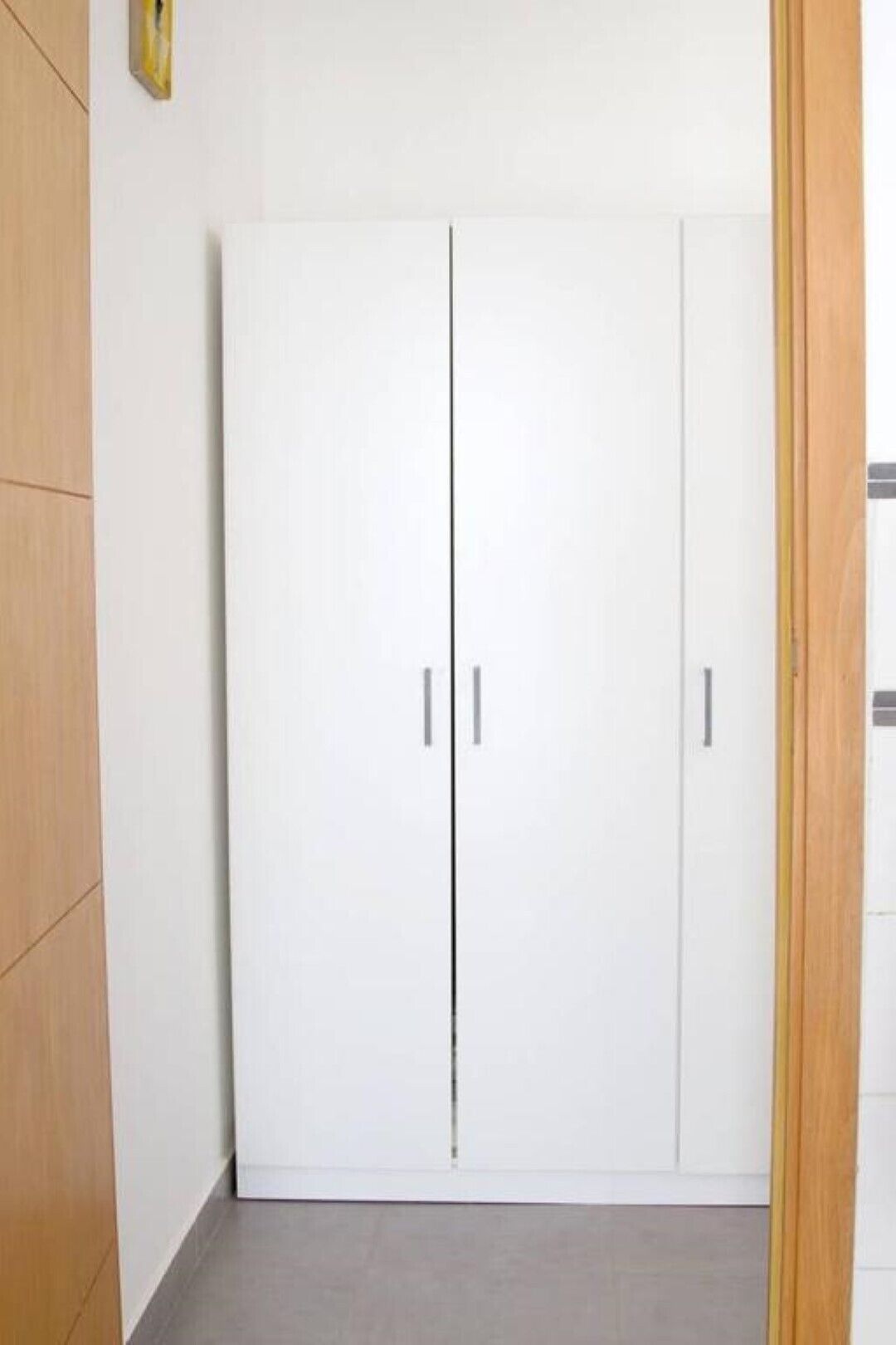 Un armario blanco con dos puertas y una manija central, frente a una pared lisa con suelo de madera.