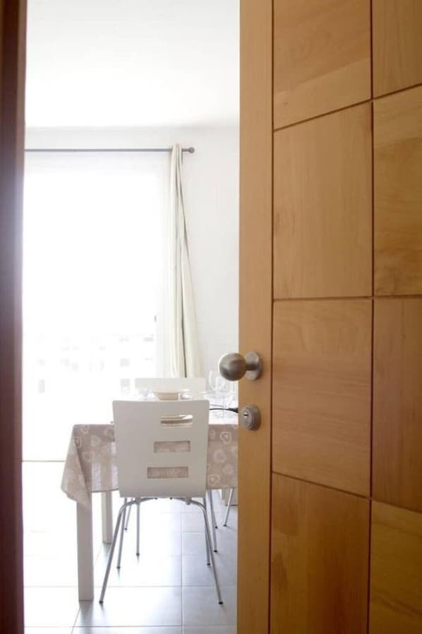 A wooden door with a round handle leading to a room with a white table and chairs, a window with curtains, and a tiled floor.