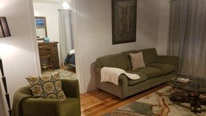 Un salon avec un canapé vert, un fauteuil à motifs, une table basse en verre et un plancher en bois.