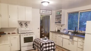 Une cuisine avec des meubles blancs, un fourneau, une table recouverte d’une nappe à carreaux et une fenêtre donnant sur des arbres.