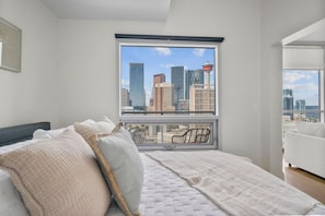 Quarto com vista para a cidade, cama com almofadas, janela com persianas e uma cadeira.