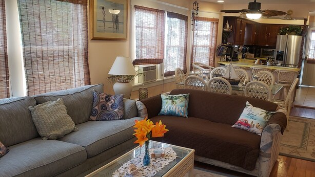 A living room with a sofa, coffee table, and decorative pillows. There is a dining area with chairs and a table. The kitchen is visible in the background with cabinets and appliances. - A Sandcrab & Jellyfish Walk Into a Sand-Bar (Ocean City)