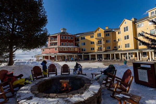 A snow-covered outdoor seating area with a fire pit, surrounded by multi-story buildings with balconies.