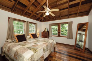 Ein Schlafzimmer mit Holzboden, einem großen Bett mit gemustertem Quilt, zwei Fenstern mit Vorhängen, einem Schminktisch und einem Spiegel.