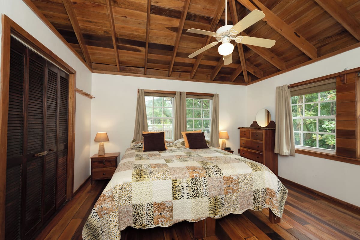 A bedroom with a wooden ceiling, a bed with a quilt, two windows, and a nightstand with a lamp.