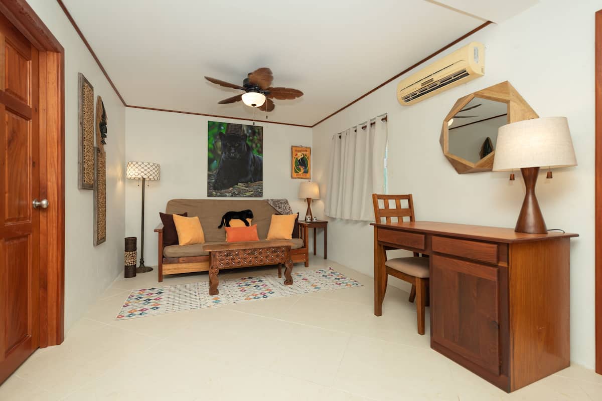 A room with a wooden desk, a sofa with cushions, a ceiling fan, a wall-mounted mirror, and a framed picture.
